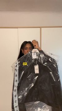 Schwarze Jacke in transparenter Verpackung mit Anhängern und Etikett. Das Material wirkt glatt. Lange Ärmel und Knöpfe sichtbar.