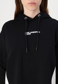 Hanorac negru cu glugă, din bumbac, având un logo frontal "Karl Lagerfeld Jeans" în alb, cordon ajustabil și o textură moale.