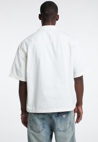 Camicia bianca a maniche corte con colletto, caratterizzata da cuciture tono su tono e una vestibilità rilassata. Abbinata a pantaloni in denim azzurro chiaro, con una toppa con logo.