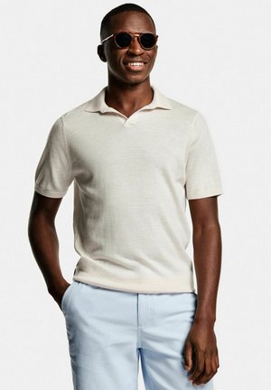 Polo shirt beige clair à manches courtes et col en V, associé à un pantalon bleu clair. Le modèle porte des lunettes de soleil rondes.