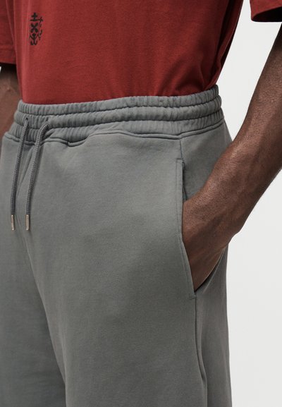 Pantalon de survêtement gris en tissu doux avec une ceinture élastique, des poches latérales et un cordon de serrage. Un t-shirt rouge foncé recouvre la ceinture.