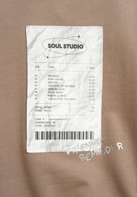 Ein Stoffaufnäher auf brauner Kleidung zeigt eine nachgemachte Quittung von "Soul Studio", die positive Handlungen ohne Preisangabe und die Worte "Freundliche Erinnerung" auflistet.
