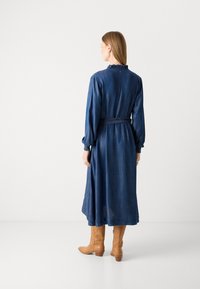 Dolga denim obleka z nabranim izrezom, dolgimi rokavi in zavezanim pasom. Krilo sega pod koleno. Oblečena s svetlimi gležnjarji.