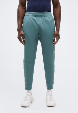 Άνδρας που φοράει τιρκουάζ παντελόνι τζόγκινγκ Nike, λευκές κάλτσες, λευκά αθλητικά παπούτσια και ναυτικό μπλε αμάνικο τοπ, στέκεται μπροστά σε απλό λευκό φόντο.