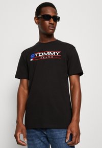 Černé bavlněné tričko s červeným a bílým nápisem "TOMMY JEANS", detailem americké vlajky, krátkými rukávy a střihem s kulatým výstřihem.