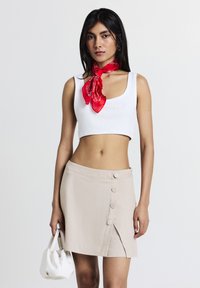 Débardeur blanc court, mini-jupe beige avec des boutons, et écharpe rouge à motifs. Tissu doux et détail fente sur la jupe. Sac à main blanc.