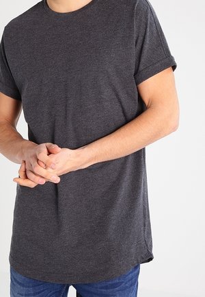 Homme portant un t-shirt gris foncé à manches courtes et un jean bleu, les mains jointes devant le torse sur un fond uni.