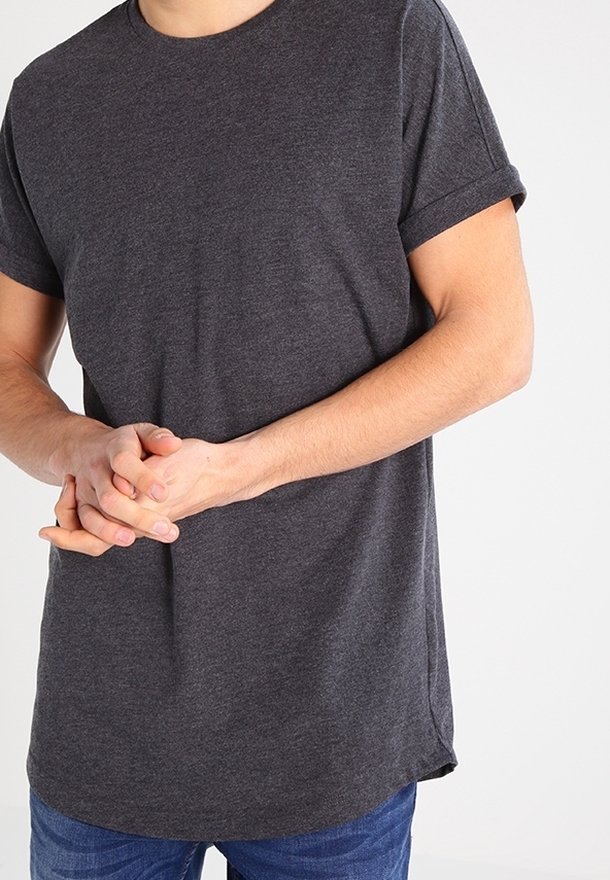 Homme portant un t-shirt gris foncé à manches courtes et un jean bleu, les mains jointes devant le torse sur un fond uni.