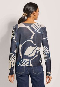 Marineblauer und cremefarbener Pullover mit floralem Design, langen Ärmeln, rundem Ausschnitt und weicher Textur. Getragen mit dunklen Denim-Hosen, Rückansicht gezeigt.