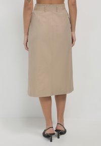 Femme debout pieds nus, portant une jupe beige mi-longue et des sandales à talons noirs, vue de dos avec un tatouage sur le bras gauche.