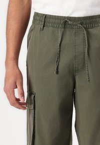 Olivgrüne Cargo-Hosen mit elastischem Bund, Kordelzug, Seitentasche und strukturiertem Stoff. Eine Hand, die auf dem Stoff ruht, wird teilweise gezeigt.