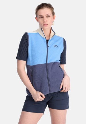 Fleece vest featuring light blue, cream, and navy panels, front zipper, and side pockets. Worn over a dark short-sleeved top and navy shorts.