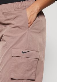 Světle růžové cargo šortky s elastickým pasem, mají boční kapsu a černé logo Nike na klopě. Materiál vypadá hladce.