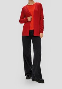Cardigan rouge en tissu doux, design ouvert à l'avant, associé à un haut orange texturé. Jean large foncé et chaussures noires massives.