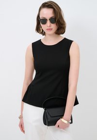 Schwarzes ärmelloses Peplum-Top aus glattem Stoff, kombiniert mit weißen Hosen. Die Accessoires umfassen eine schwarze Handtasche und große goldene Ohrringe.