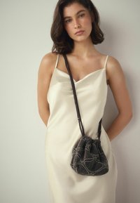 Geantă tip bucket neagră cu șnur, cu un model argintiu, având o țesătură texturată și o curea lungă, închisă la culoare. Pur tată peste o rochie de culoare crem.