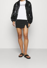 Svart, blank bomberjacka med elastiska manschetter, vit T-shirt med logotyp, svart minikjol med sidoklyfta och svarta slidesandaler.