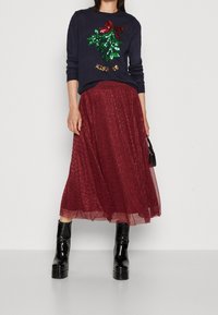 Marineblauwe trui met een sequined maretakontwerp en de tekst "KISS ME", gecombineerd met een bordeauxrood, glinsterende, enkellange tule rok en zwarte laarzen.