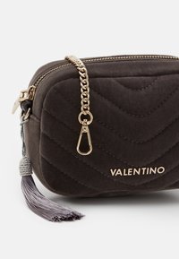 Tmavě šedá prošívaná semišová taška se zlatým kování, popruhem z propojeného řetězu a šedým střapcem. Disponuje zipovým uzávěrem a logem.