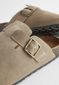 Sandales en suede marron avec deux sangles ajustables, chacune dotée d'une boucle en laiton. La semelle intérieure texturée contraste avec la semelle extérieure rigide en caoutchouc noir.