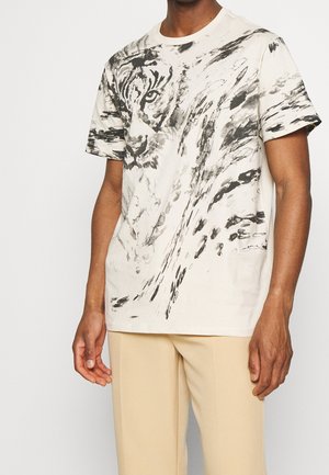 Mann trägt off-white T-Shirt mit schwarzem und grauem abstraktem Tigerkopfprint, kombiniert mit beigen Hosen, stehend vor weißem Hintergrund.