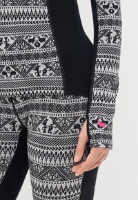 Lämpimät paidat ja housut mustasta ja harmaasta neulotusta kankaasta, joissa on geometrisia kuvioita. Piirteet include vaaleanpunainen logo-aksentti ja teksturoidut ribbialueet saumoissa.