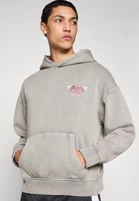 Ανοιχτό γκρι φούτερ με κουκούλα, εμπρόσθια τσέπη, ριμπ μανσέτες και ζώνη μέσης. Διαθέτει κόκκινο και μπλε λογότυπο σε γραφική απεικόνιση στο στήθος. Άνετη, καθημερινή εφαρμογή.