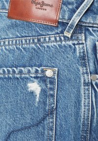 Světle modré džíny z denimu s opotřebovaným vzhledem, stříbrné kovové doplňky, viditelné švy a hnědá kožená etiketa na pásku.