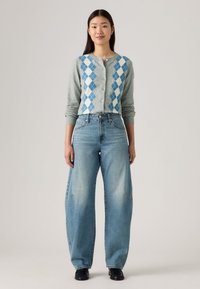 Cardigan gris clair avec un motif argyle bleu et blanc, fermeture à boutons, associé à un jean bleu ample et des chaussures noires.