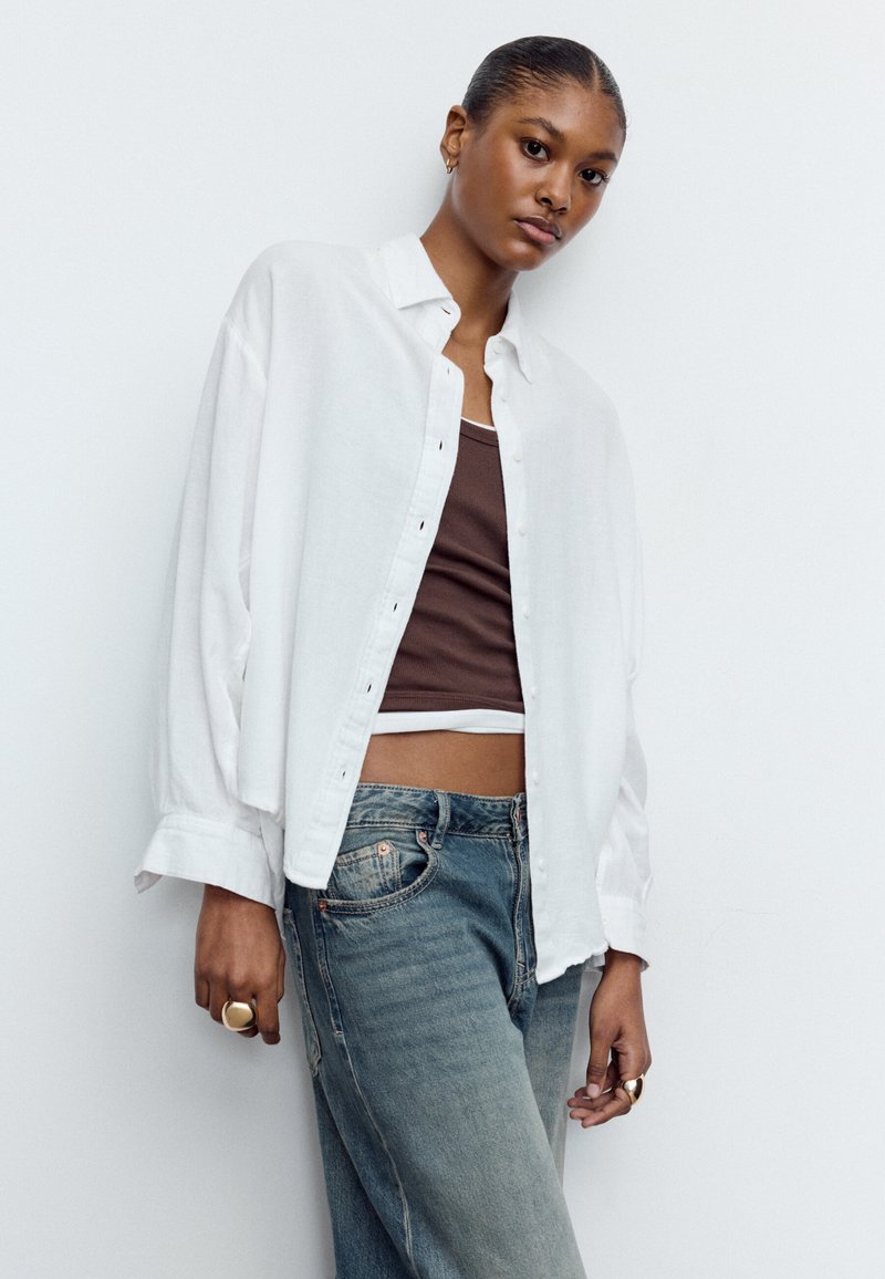 Ung kvinde med glat hår trukket tilbage iført en åben hvid skjorte over en brun crop top og blå jeans, stående mod en ensartet hvid væg.