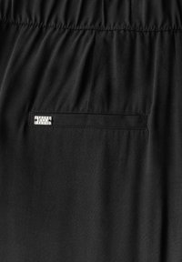 Tissu noir au toucher lisse avec une poche discrète et une plaque logo en métal argenté. La taille élastique ajoute à son design.