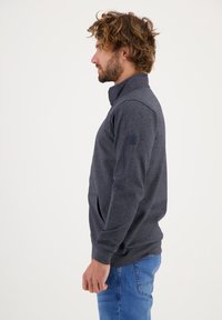 Pullover grigio in pile con colletto alto, maniche lunghe, tasche laterali e patch con logo sulla manica sinistra, abbinato a jeans blu.