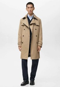 Beige trenchcoat met een dubbelzijdig ontwerp, grote kraag en knoopdetails, gedragen over een marineblauw vest en een blauwe shirt. Donkere broek en schoenen.