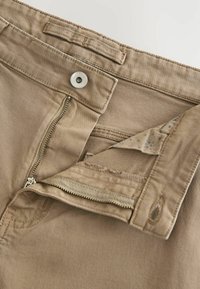 Pantalon en coton beige avec fermeture éclair et bouton à l'avant, en tissu texturé et avec une doublure intérieure à motifs visible à l'ouverture.