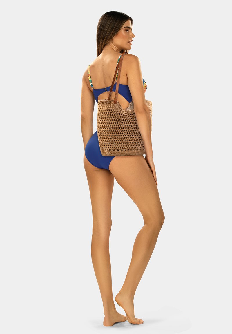 Spleciona torba w kolorze beżowym z brązowymi skórzanymi paskami, posiadająca otwarty wzór. Modelka w niebieskim kostiumie kąpielowym pozuje tyłem.