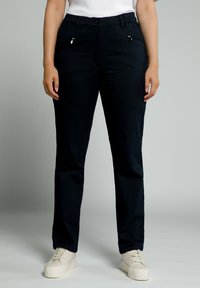 Pantalones negros con corte slim, que presentan bolsillos con cremallera en la parte delantera y una textura suave, combinados con zapatillas blancas.