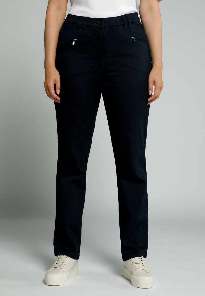 Pantalones negros con corte slim, que presentan bolsillos con cremallera en la parte delantera y una textura suave, combinados con zapatillas blancas.