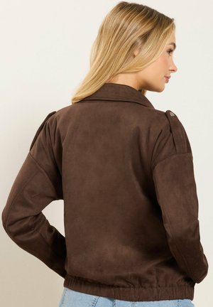 Kvinde med langt blondt hår iført en brun ruskindsjakke med skulderepåletter, vist bagfra og i sideprofil.