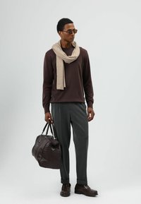 Chemise marron à manches longues, écharpe beige clair, pantalon plissé gris, chaussures en cuir marron et sac de voyage en cuir marron foncé.