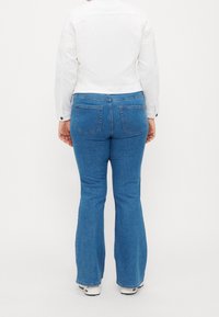 Person sedd bakifrån som bär en vit jacka, blårandiga bootcut jeans och vita sneakers mot en helt vit bakgrund.