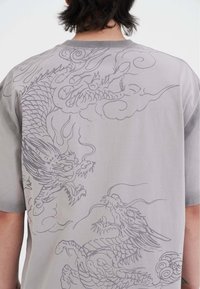 Camiseta gris con una gran impresión detallada de un dragón en gris oscuro en la parte posterior, que muestra nubes en remolino y escamas intrincadas. Mangas cortas.