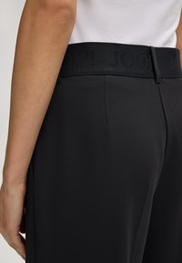 Zwarte broek met een gladde textuur, voorzien van een verhoogde donkere logo-band en een getailleerde pasvorm. Zijzakken zijn zichtbaar op de heupen.