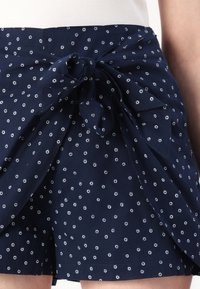 Tmavě modré šortky s mašlí v pase, zdobené rozptýleným vzorem bílých teček a vyrobené z hladké, lehké látky.
