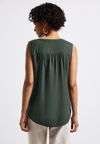 Bezrukává zelená blůza s nařaseným detailem na zadní straně, hladkou texturou a zakřiveným spodním lemem; kombinována se světlými kalhotami.