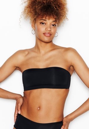 Giovane donna con capelli ricci che indossa un top bikini bandeau nero e slip abbinati, posando con le mani sui fianchi su sfondo bianco.