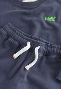 Sudadera de color azul marino y pantalones a juego con cintura elástica y cordones blancos, con el logotipo verde de SuperDry en el pecho de la sudadera.