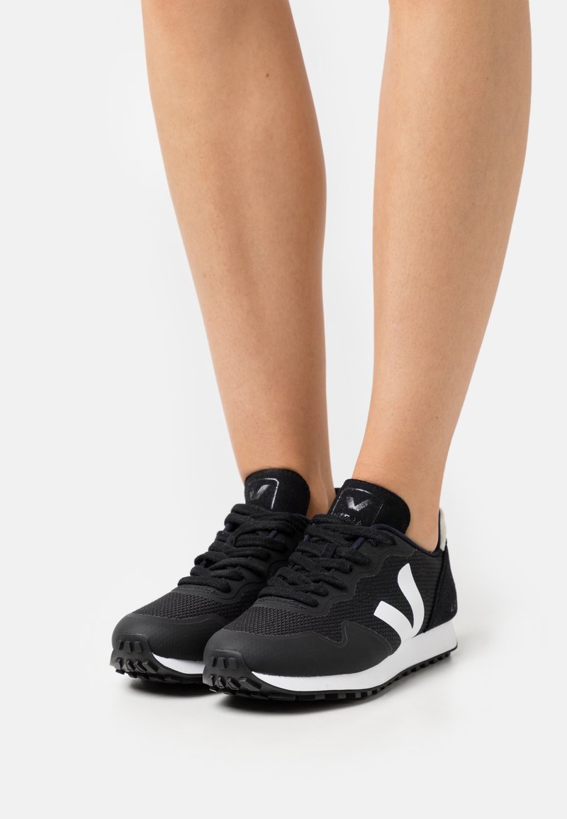 Chaussures de sport noires avec une tige en mesh, des accents blancs, une semelle en caoutchouc texturée et une étiquette de marque contrastante au talon.