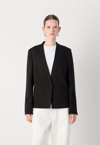 Blazer nero con design a scollo a V, chiusura a un bottone, tasche frontali e tessuto testurizzato, indossato sopra una maglia bianca e pantaloni chiari.