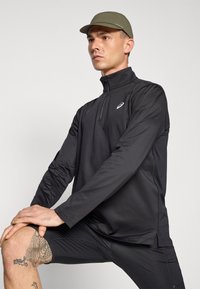 Svart langermet treningstopp med glidelås i kragen og reflekterende logo, kombinert med svarte shorts. Modell med grønn caps.