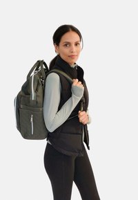 Mochila acolchada verde con acentos negros, bolsillos con cremallera y correas acolchadas. El modelo lleva un chaleco negro y una camiseta de manga larga azul claro.